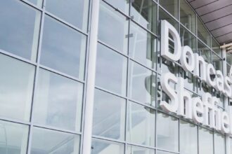 An up-close image of the outside of the entrance of Doncaster Sheffield Airport. The building is made of glass panels which reflect the sky and clouds. Viewable is white lettering that says 'Doncaster Sheffield'.