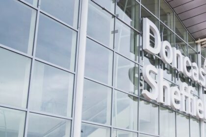 An up-close image of the outside of the entrance of Doncaster Sheffield Airport. The building is made of glass panels which reflect the sky and clouds. Viewable is white lettering that says 'Doncaster Sheffield'.