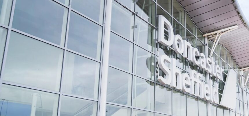 An up-close image of the outside of the entrance of Doncaster Sheffield Airport. The building is made of glass panels which reflect the sky and clouds. Viewable is white lettering that says 'Doncaster Sheffield'.