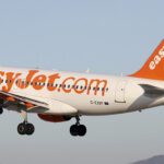 An easyJet Airbus A319 mid-flight with a blue sky in the background. The tips of two mountains can be seen in the distance.