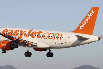 An easyJet Airbus A319 mid-flight with a blue sky in the background. The tips of two mountains can be seen in the distance.