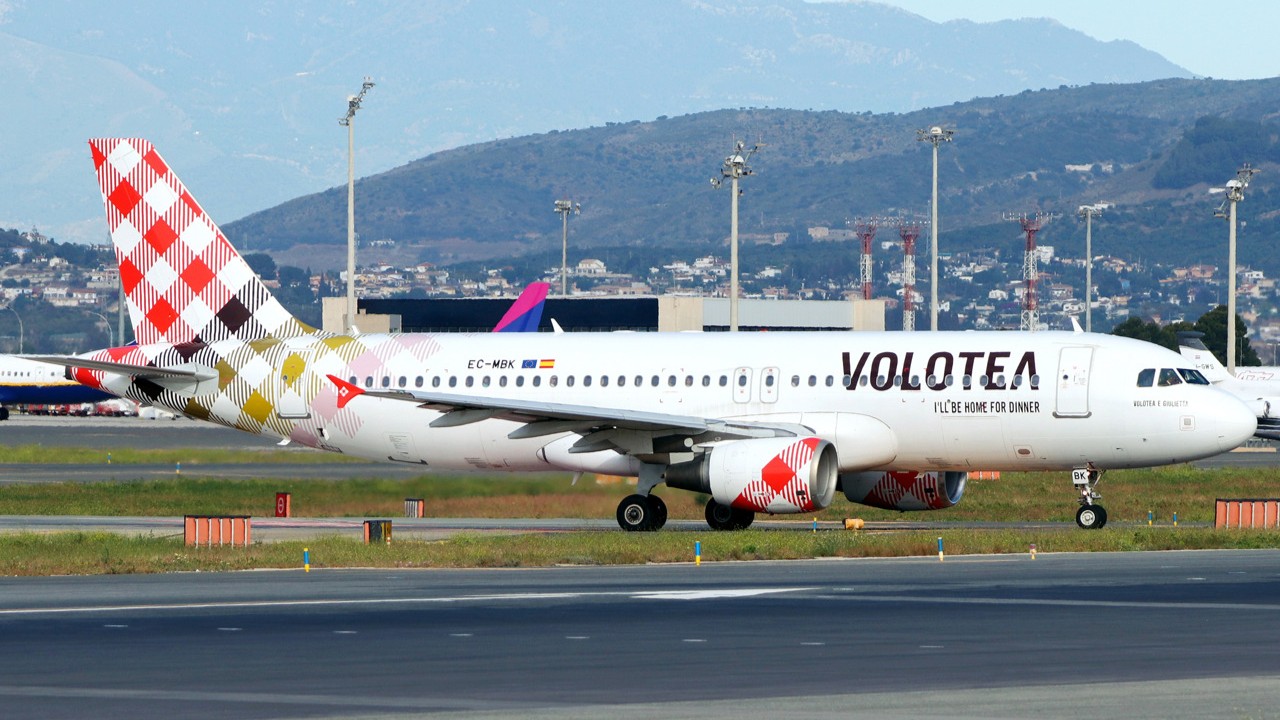 Volotea Airbus A320-200 EC-MBK taxiing at an airport, with its red and white check-pattern tail livery and hills in the background.