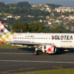 Volotea Airbus A319-100 registered EC-NHP on a runway in daylight, with a wooded hillside and houses behind it