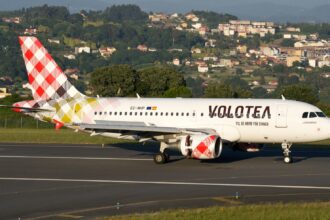 Volotea Airbus A319-100 registered EC-NHP on a runway in daylight, with a wooded hillside and houses behind it