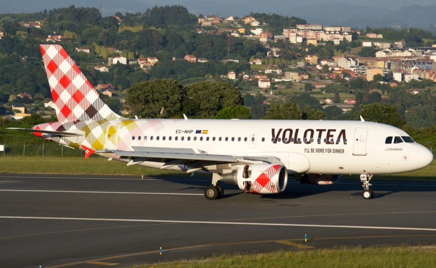 Volotea Airbus A319-100 registered EC-NHP on a runway in daylight, with a wooded hillside and houses behind it