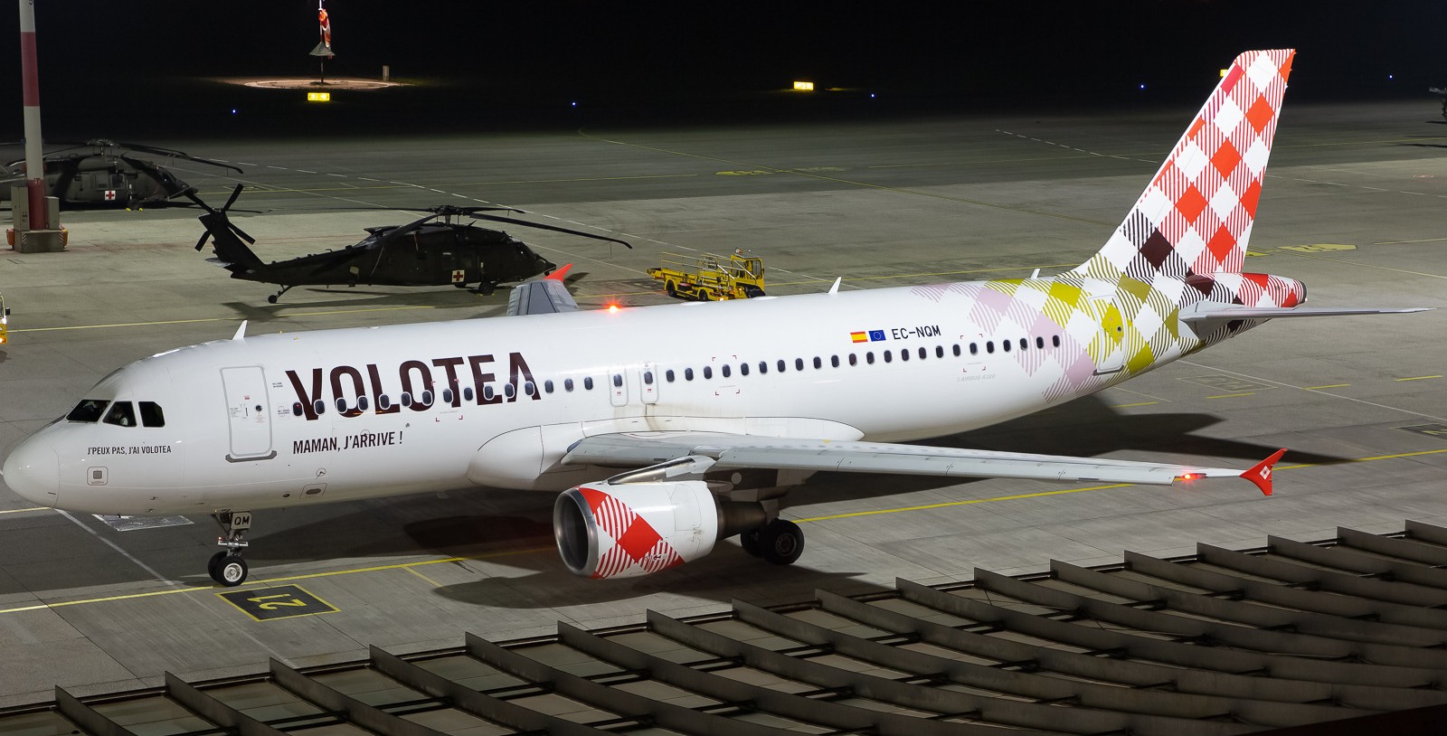 Volotea Airbus A320-200 registered EC-NQM taxiing on a floodlit apron at night, with two dark helicopters parked behind it