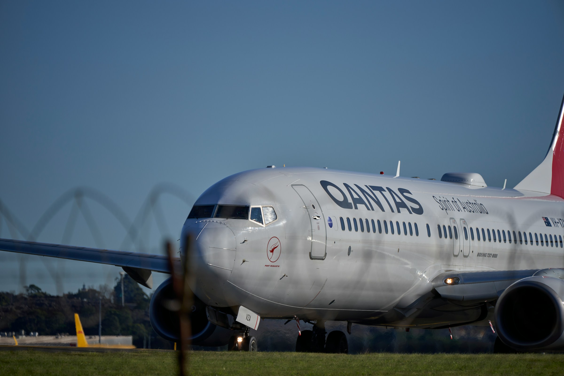 A Qantas plane on an airport runway with green fields around it and a bright blue sky in the background.