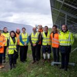 A group of people in high-vis jackets stand in a muddy field next to large solar panels.