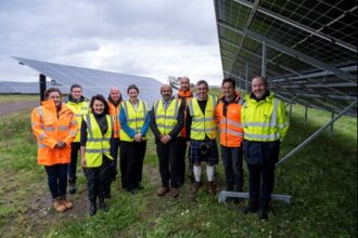 A group of people in high-vis jackets stand in a muddy field next to large solar panels.