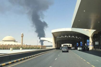 Vehicles drive along the road outside King Khalid International Airport, with a distinctive domed building and a large plume of dark smoke visible in the background.