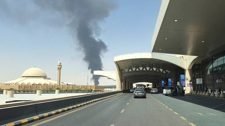 Vehicles drive along the road outside King Khalid International Airport, with a distinctive domed building and a large plume of dark smoke visible in the background.