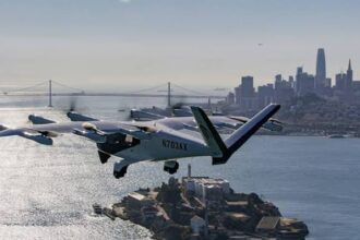 An Archer electric aircraft with tail number N703AX flies over Alcatraz Island with the San Francisco skyline and Bay Bridge visible in the background.