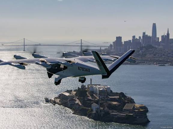 An Archer electric aircraft with tail number N703AX flies over Alcatraz Island with the San Francisco skyline and Bay Bridge visible in the background.