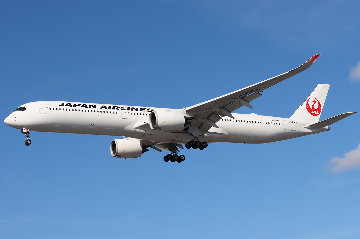 A Japan Airlines Airbus A350-1041 in the air, with its landing gear extended against a blue sky with partial cloud cover.