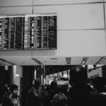 A black and white image depicting a group of passengers looking at an airport departure board.