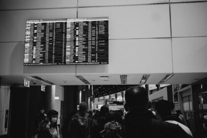 A black and white image depicting a group of passengers looking at an airport departure board.