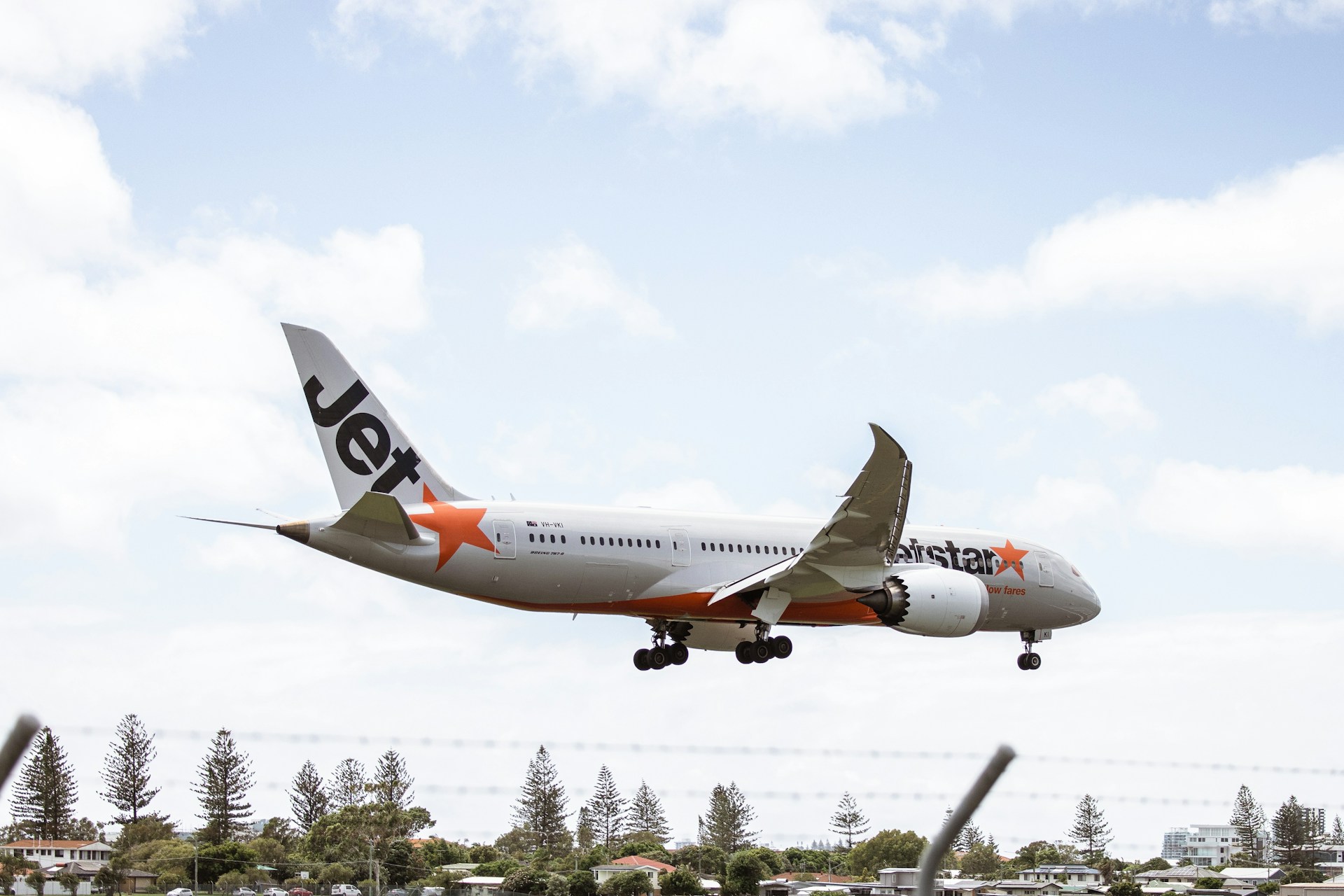 A Jetstar flight is pictured landing onto an airport runway, with its landing gear extended against a blue cloudy sky and trees in the background.