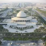 An ariel shot of King Fahd International Airport in Dammam, Saudi Arabia. A white building is at the centre of the area with a dome-shaped roof, its surrounded by areas of foliage and roads that run past other buildings.