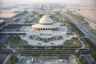 An ariel shot of King Fahd International Airport in Dammam, Saudi Arabia. A white building is at the centre of the area with a dome-shaped roof, its surrounded by areas of foliage and roads that run past other buildings.