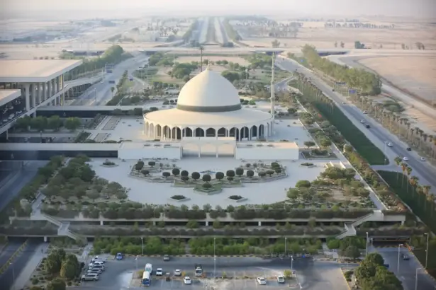 An ariel shot of King Fahd International Airport in Dammam, Saudi Arabia. A white building is at the centre of the area with a dome-shaped roof, its surrounded by areas of foliage and roads that run past other buildings.