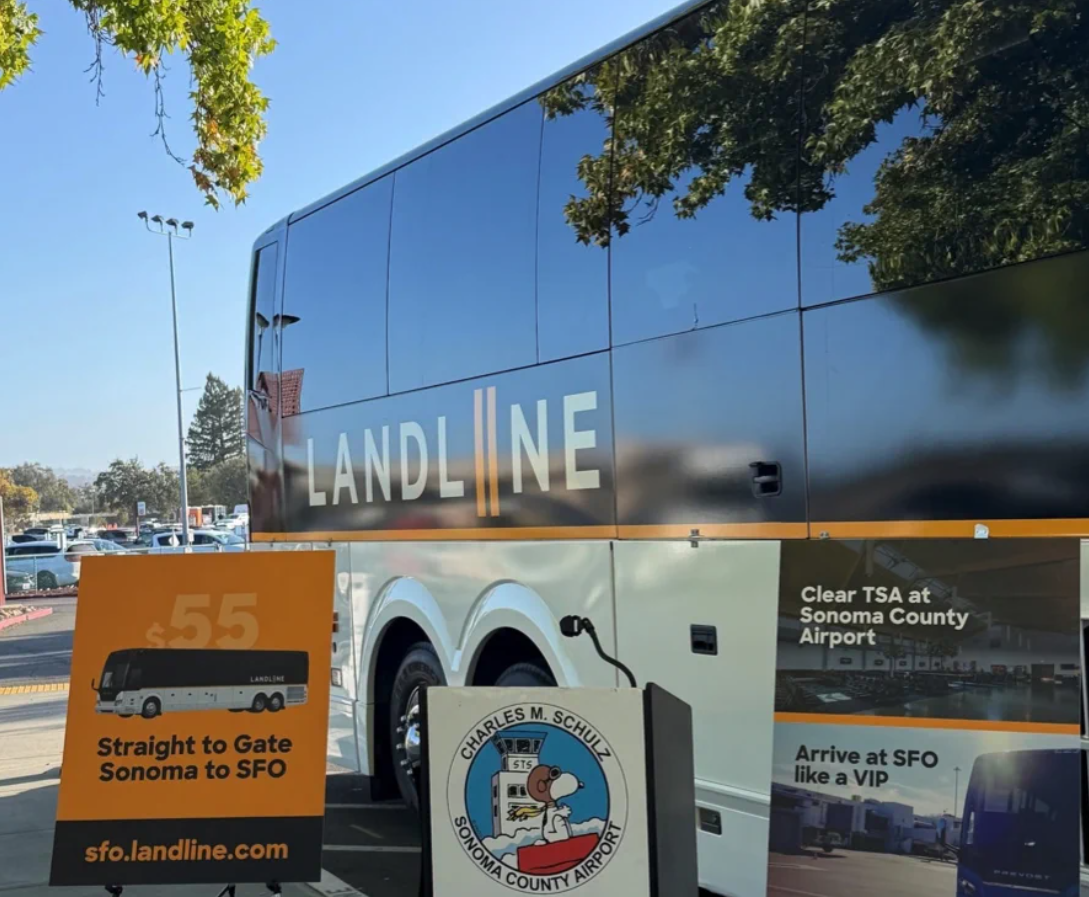 A Landline bus parked near a sign advertising a $55 shuttle service from Sonoma County Airport to SFO, featuring the Charles M. Schulz airport logo.