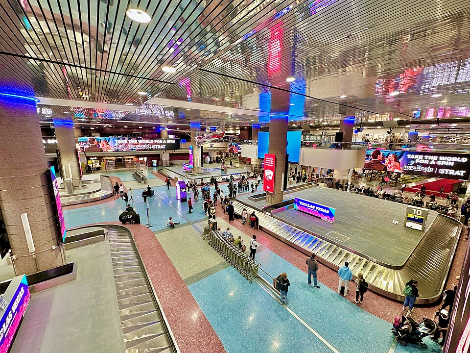 The baggage claim area in Las Vegas Airport. Multiple baggage belts can be seen in the centre of the hall with people surrounding one of them. Bright signs are on the walls of the hall.