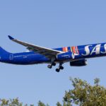 A dark blue SAS Airbus A330-343 during take off, with its landing gear extended above trees against a clear blue sky.