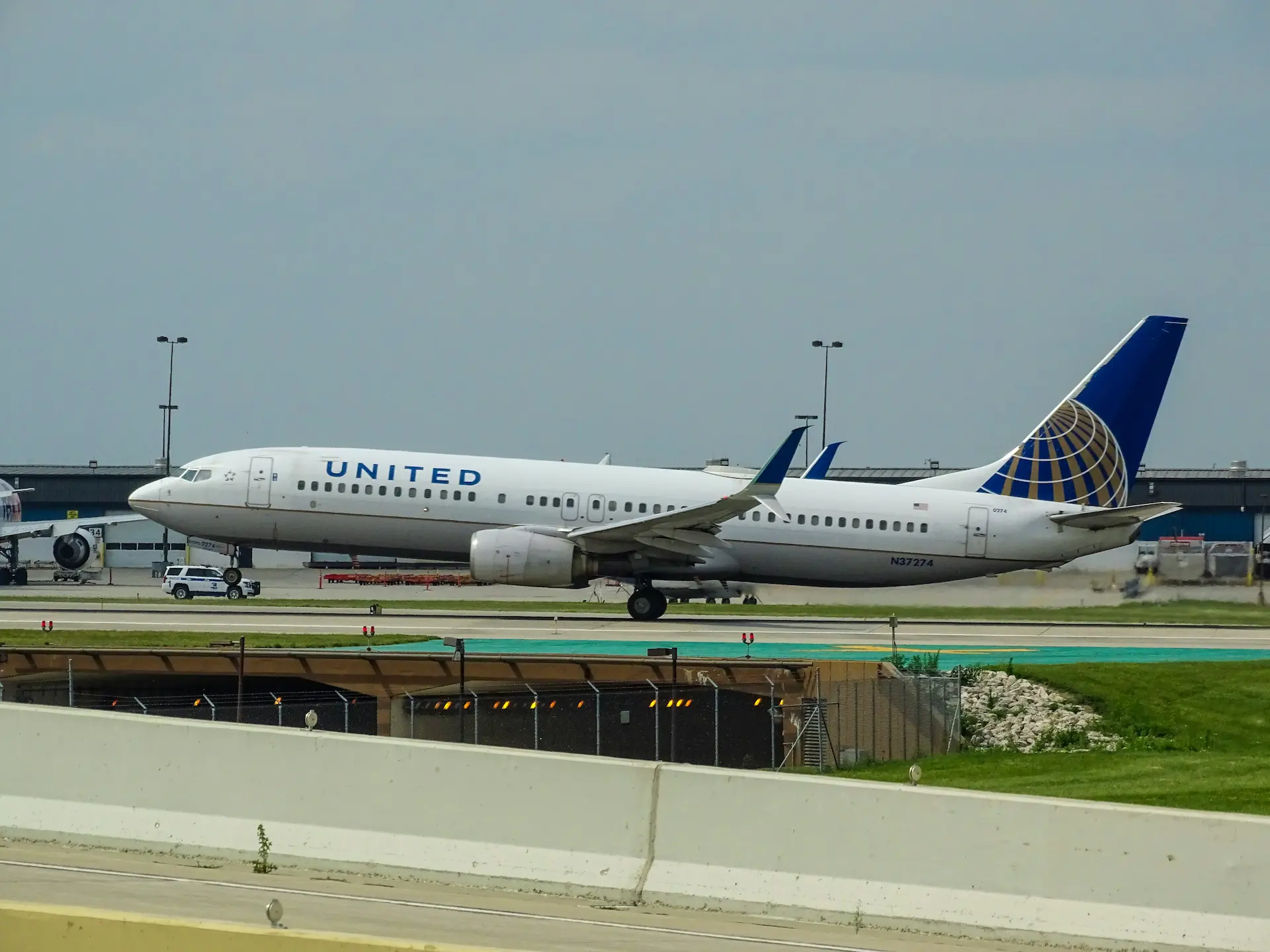United Airline Boeing 737 (N37274) taxiing along an airport runway under an overcast sky, with a patrol vehicle and airport buildings behind it.