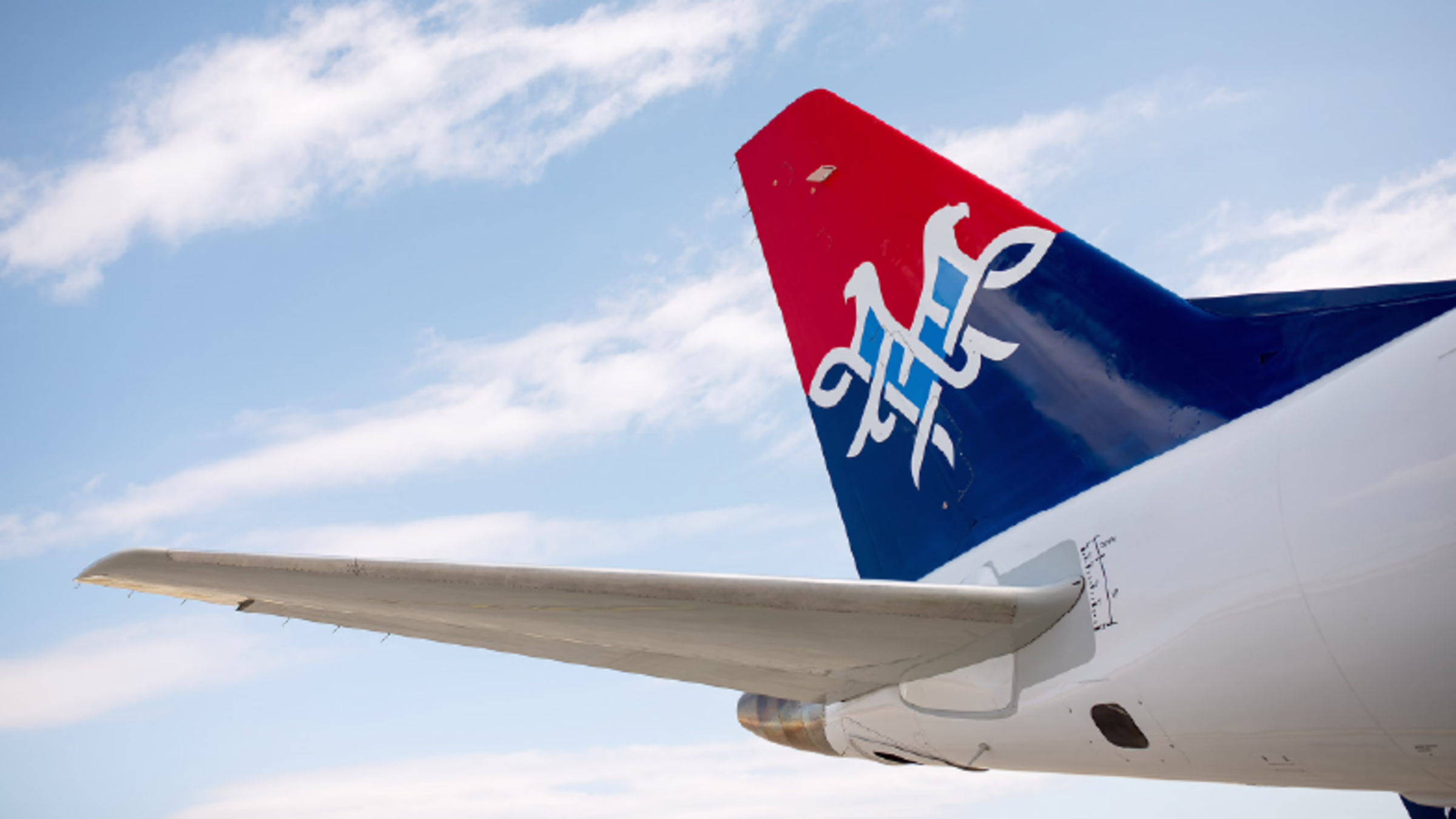 Photo of a Air Serbia wing in the right hand corner in front of a blue sky and clouds background