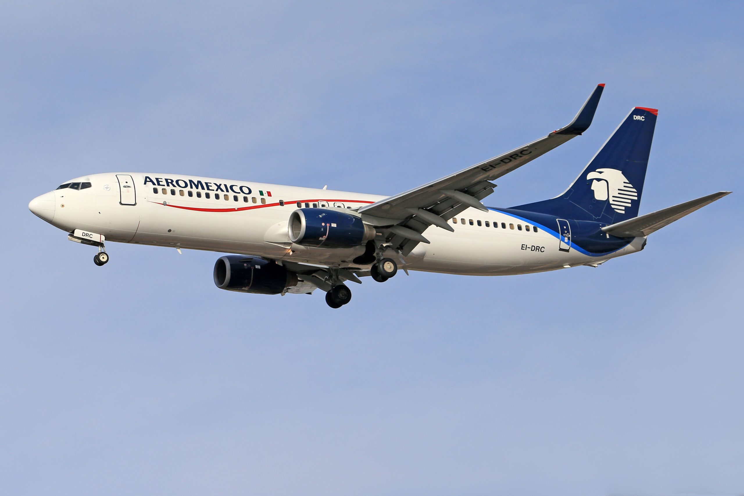 Photograph shows an Aeromexico passenger jet, likely Boeing 737, flying against a clear blue sky with landing gear extended. White fuselage, dark blue engines and tail, red-green accent stripe, and airline eagle logo identify aircraft during approach or landing.