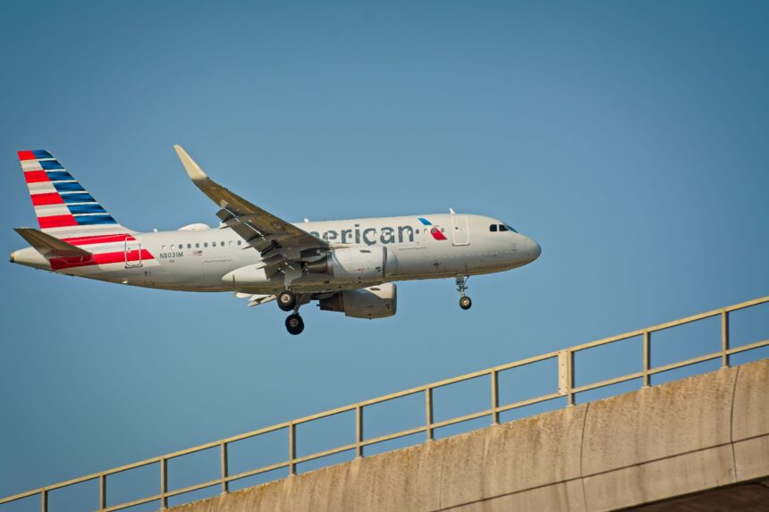 A large American Airlines airplane soars above a bridge, showcasing its wingspan against a clear blue sky.