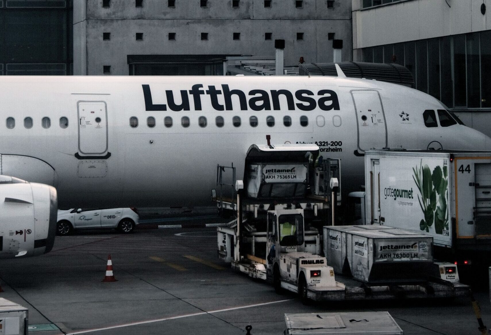 A Lufthansa airplane parked at an airport terminal with a concrete wall and ground support equipment nearby.