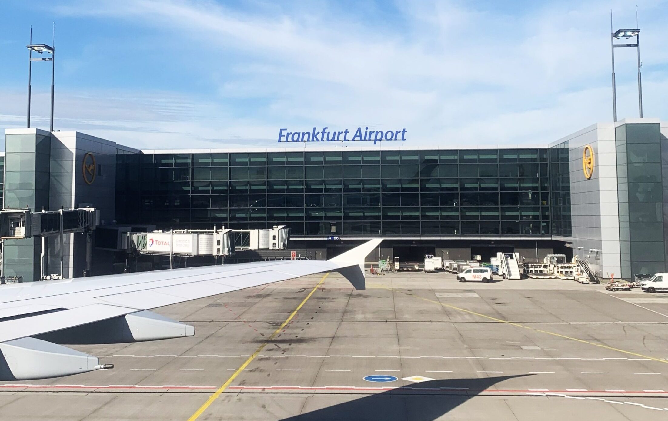 View from an airplane window showing the Frankfurt Airport terminal building with a large glass facade, jet bridges, and tarmac under a blue sky.