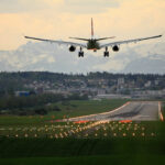 A plane is approaching a runway for landing with its landing gear extended. In the background, a mountainous region can be seen and the runway is clearly surrounded by green grass.