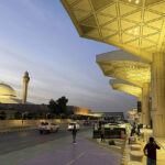 The exterior of Riyadh's King Khalid International Airport. The doors are set behind large pillars lit by warm lights. Cars and people can be seen outside of the airport's entrance.