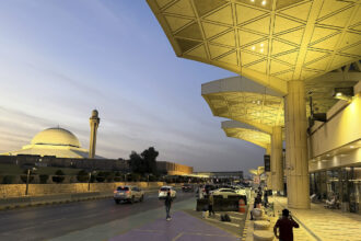 The exterior of Riyadh's King Khalid International Airport. The doors are set behind large pillars lit by warm lights. Cars and people can be seen outside of the airport's entrance.
