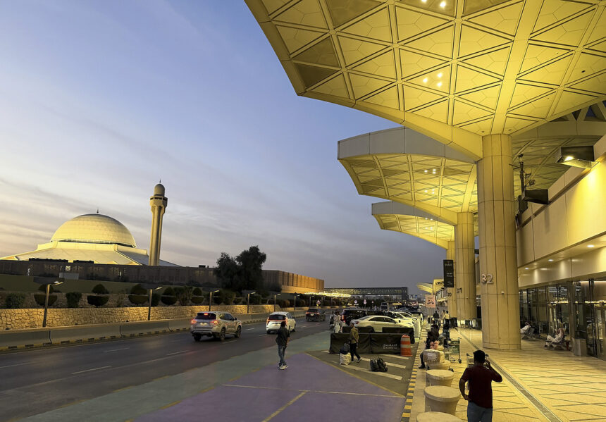 The exterior of Riyadh's King Khalid International Airport. The doors are set behind large pillars lit by warm lights. Cars and people can be seen outside of the airport's entrance.