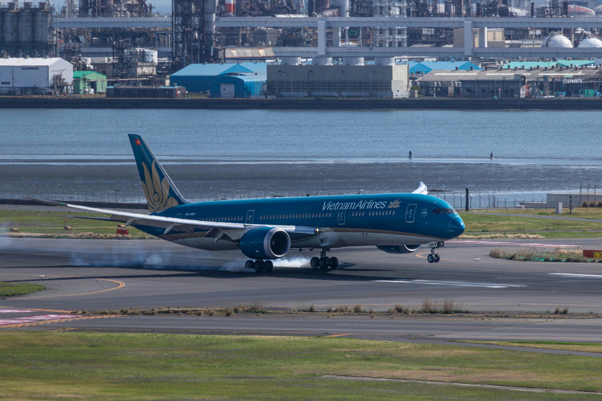 A Vietnam Airline Boeing 787-9 landing on a runway with tyre smoke, with an industrial waterfront behind.
