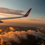A Qantas aircraft wing is pictured from the view of a passenger window mid-flight over the clouds. A blue and red sunset engulfs the sky.