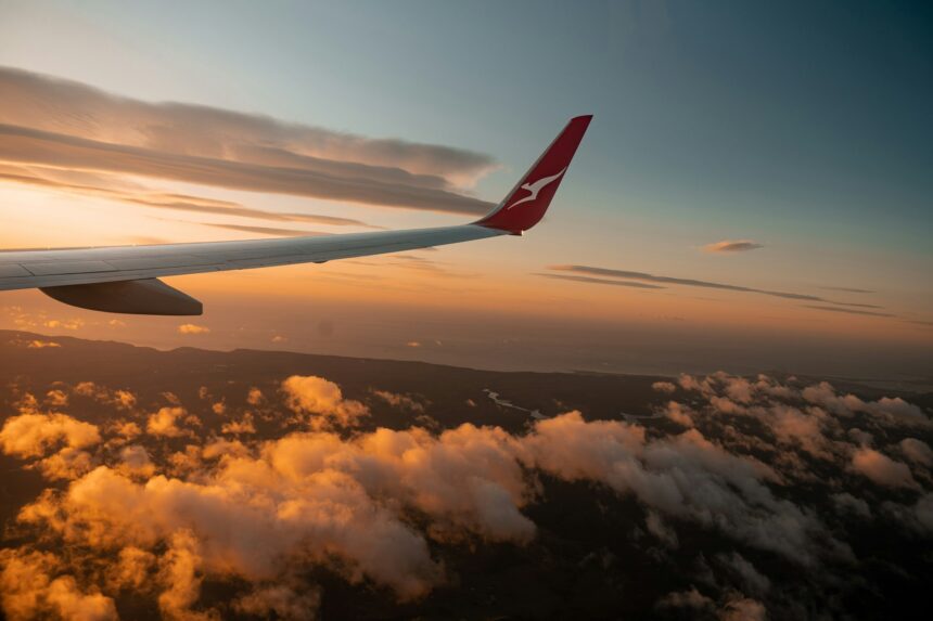 A Qantas aircraft wing is pictured from the view of a passenger window mid-flight over the clouds. A blue and red sunset engulfs the sky.