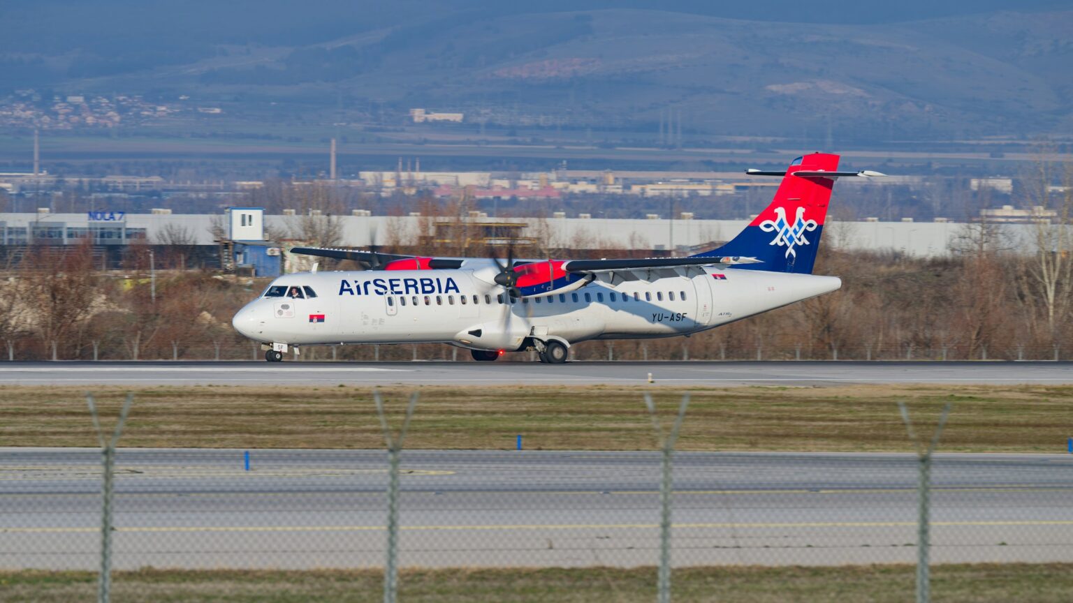 Air Serbia plane pictured landing on an airport runway with grass and trees in the background.