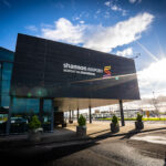 Image shows the exterior of Shannon Airport in Ireland with logo, passengers and sunshine