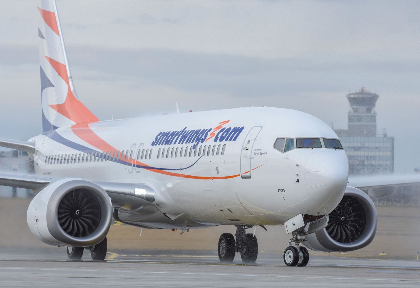 Smartwings Boeing 737 MAX 8 taxiing on a wet runway, with an airport control tower visible in the background.