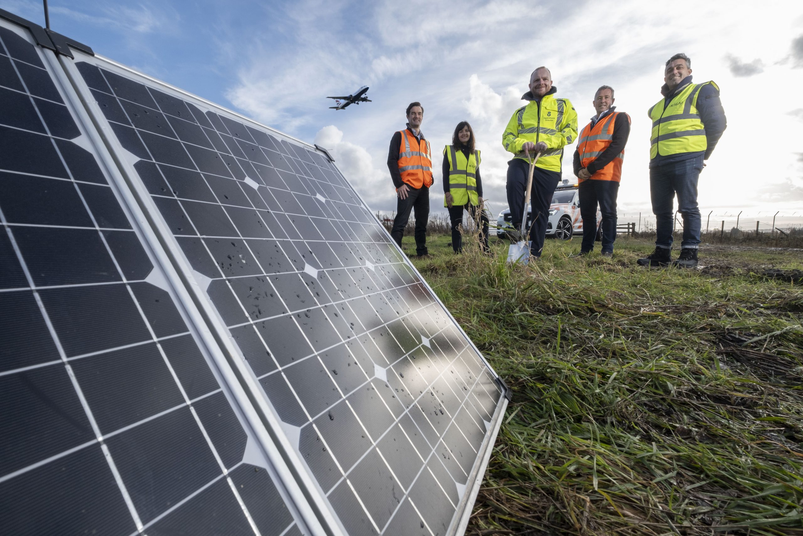 Five people are stood in a field wearing high-vis jackets, one of whom is holding a shovel. They are stood behind a large solar panel and a plane can be seen mid-flight in the background.