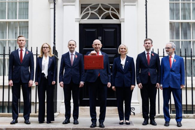 The Chancellor of the Exchequer John Healey stood outside Downing Street along with other British government officials in a line. Healey stands with one arm extended holding the red budget box.
