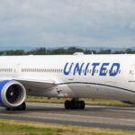 An United Airlines B787 taxiing along the runway. The concrete runway is surrounded by areas of green grass, and there is a grey sky in the background.