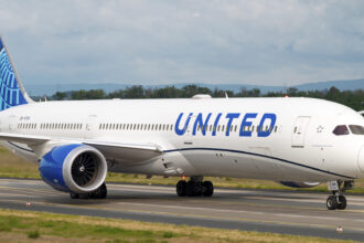 An United Airlines B787 taxiing along the runway. The concrete runway is surrounded by areas of green grass, and there is a grey sky in the background.