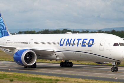 An United Airlines B787 taxiing along the runway. The concrete runway is surrounded by areas of green grass, and there is a grey sky in the background.
