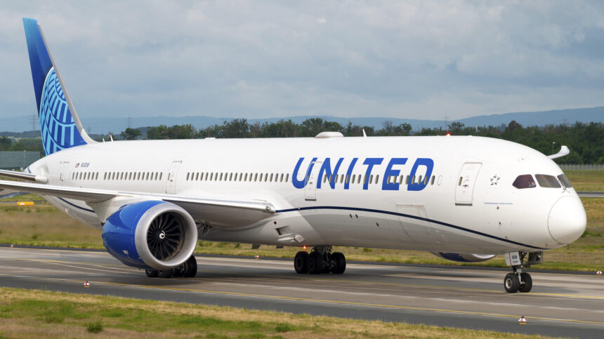 An United Airlines B787 taxiing along the runway. The concrete runway is surrounded by areas of green grass, and there is a grey sky in the background.