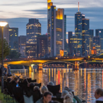 Frankfurt skyline at dusk viewed from the banks of the Main River, with illuminated skyscrapers, city lights reflecting on the water, and people walking and sitting along the riverside promenade.